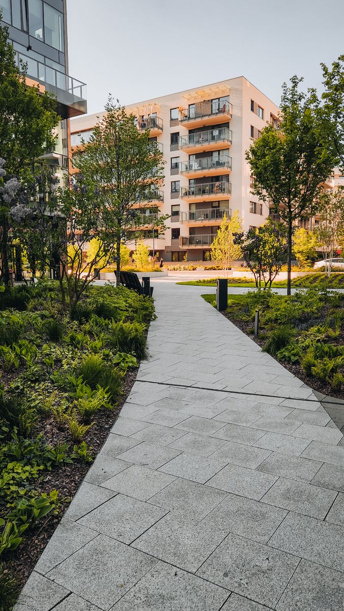 A tranquil modern residential pathway surrounded by trees and greenery with modern apartments in the background.